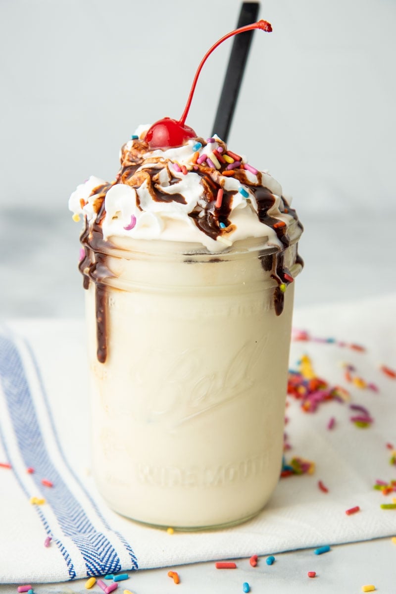 A mason jar is filled with vanilla ice cream. The ice cream is topped with whipped cream, sprinkles, chocolate sauce, and a cherry.