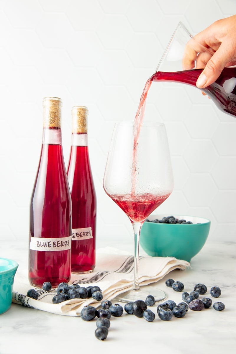 A hand pours homemade fruit wine from a carafe into a stemmed wine glass.