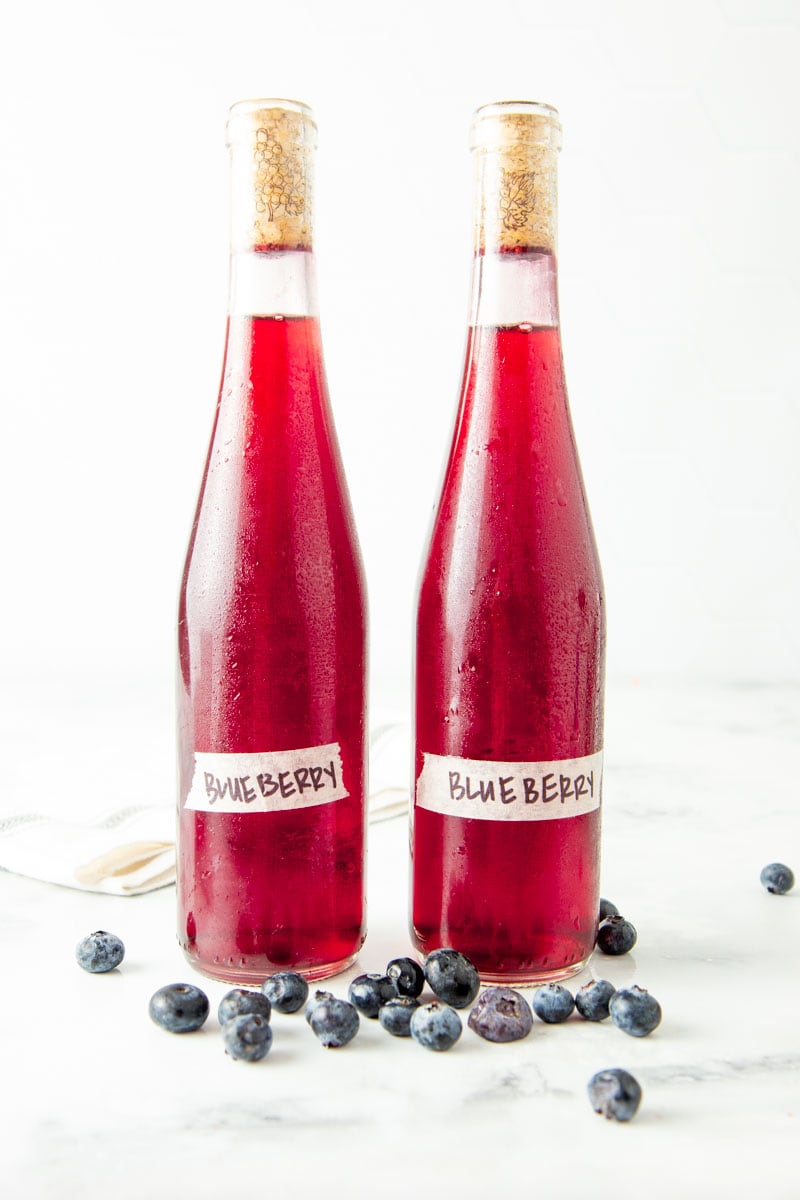 Two chilled bottles of blueberry wine stand on a white marble countertop surrounded by fresh blueberries.