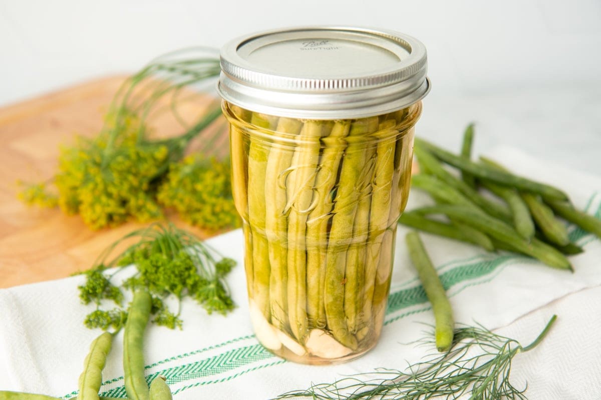 Canned dilly beans in a pint jar, with fresh green beans and dill surrounding the jar