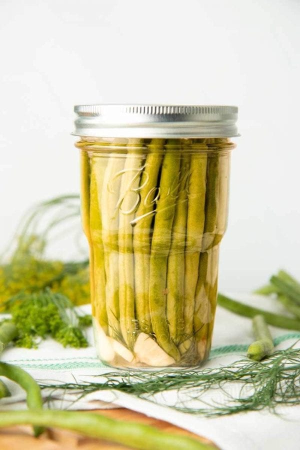 A jar of canned dilly beans sits on a dish towel surrounded by fresh dill and additional raw beans