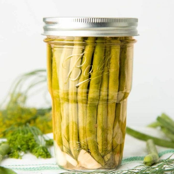 A jar of canned dilly beans sits on a dish towel surrounded by fresh dill and additional raw beans