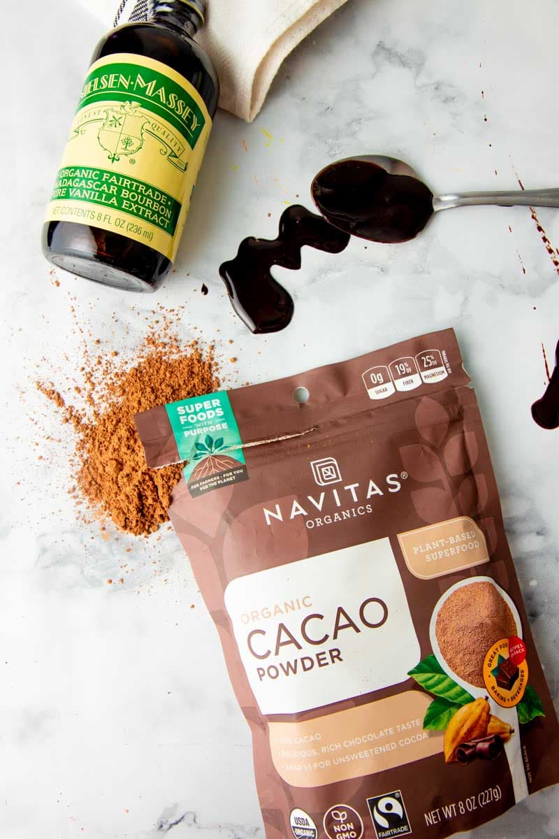 Overhead of fairtrade ingredients such as Nielsen-Massey vanilla extract and Navitas Organics cacao powder on a white marble countertop.