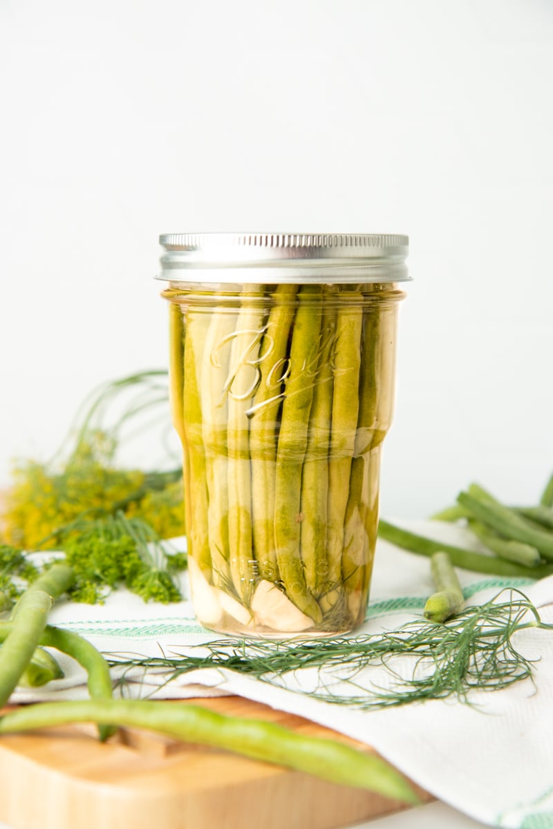 A jar of pickled dilly beans sits on a tea towel on top of a wooden cutting board. Fresh dill and additional beans are around the jar.