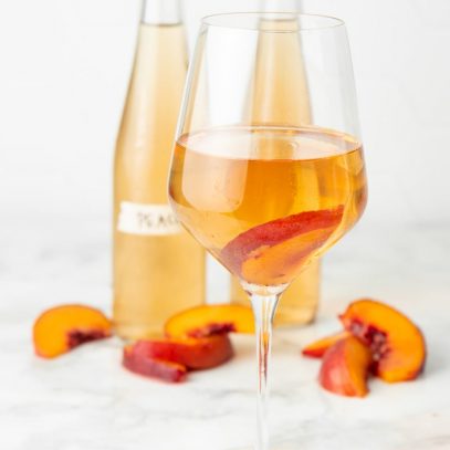 A glass of wine stands in front of two bottles and fresh peach slices.