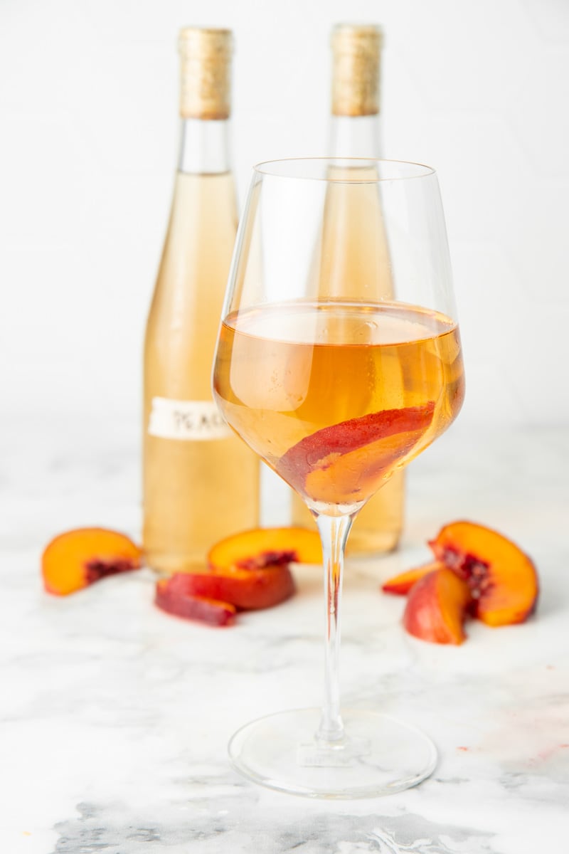A glass of wine stands in front of two bottles and fresh peach slices.
