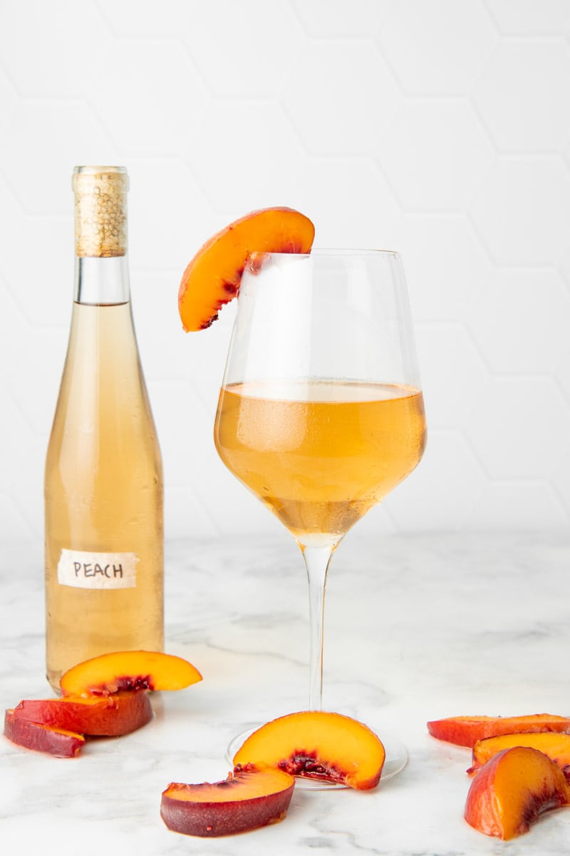 A chilled glass of homemade country wine stands with fresh peach slices around it, and a full bottle behind it.