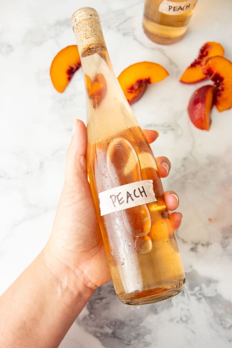 A hand holds a bottle of homemade fruit wine with a masking tape label that reads "Peach."