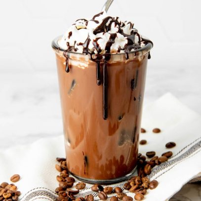 Iced mocha in a glass garnished with whipped cream and mocha syrup drizzle sits on a kitchen linen with coffee beans.
