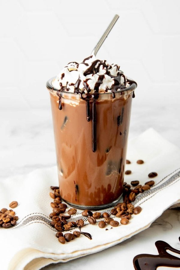 Iced mocha in a glass garnished with whipped cream and mocha syrup drizzle sits on a kitchen linen with coffee beans.