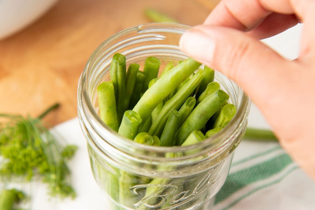 A hand nestles green beans into a glass Ball jar