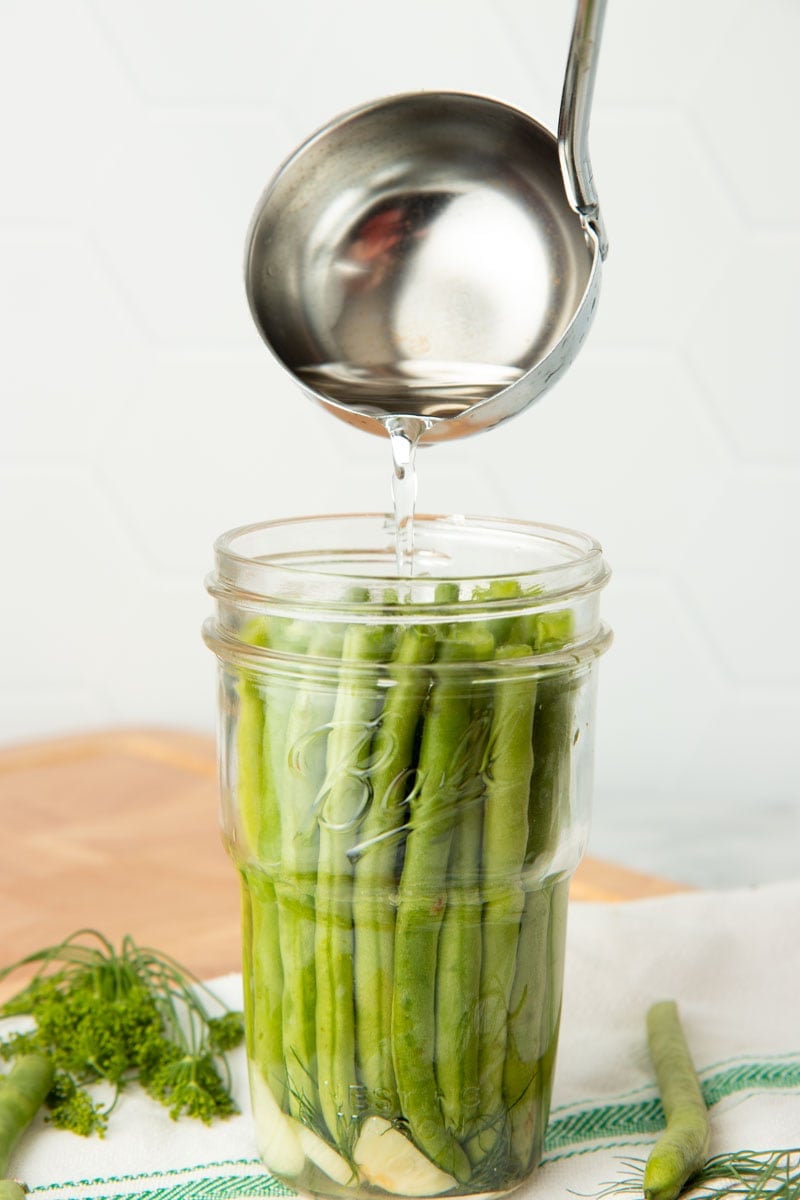 A ladle pours brine over beans packed into a glass jar