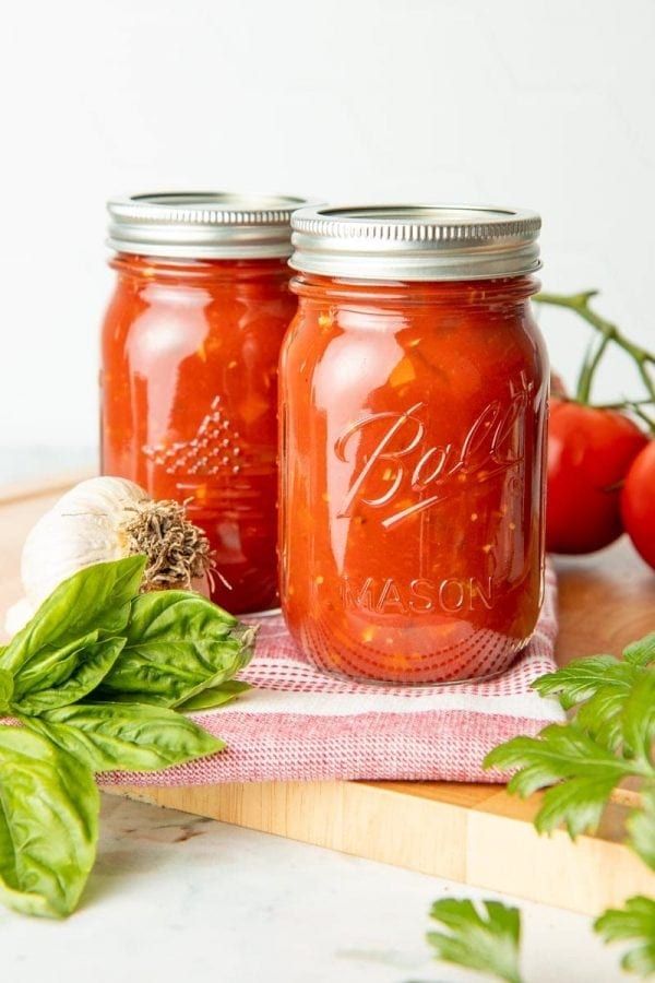 Two full canning jars of spaghetti sauce surrounded by fresh garlic, tomatoes, and herbs.
