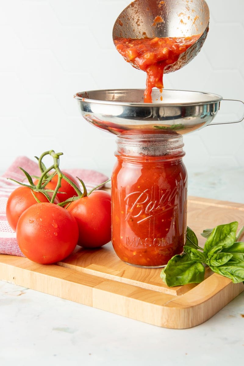A ladle pours spaghetti sauce through a funnel and into a glass canning jar.