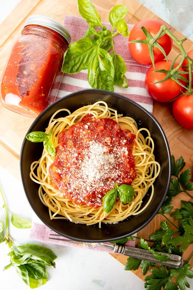 A black bowl is filled with spaghetti, sauce, and basil. The bowl is surrounded by fresh herbs, fresh tomatoes, and a jar of sauce.