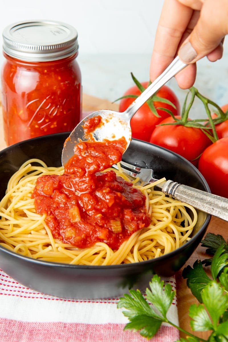 A hand spoons spaghetti sauce over a bowl of spaghetti. A full jar of canned spaghetti sauce and some fresh tomatoes sit behind the bowl.