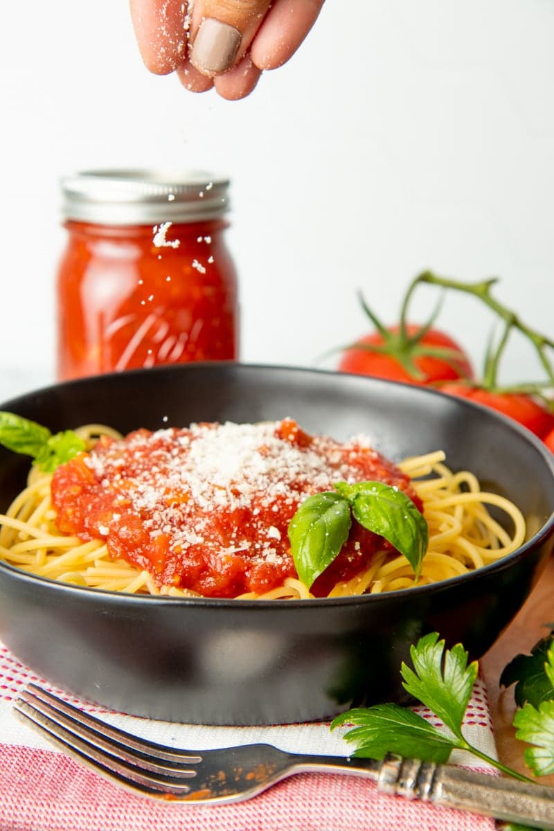 A hand sprinkles Parmesan over a bowl of spaghetti with sauce and basil