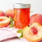 Peach jam sits in a half pint jar, surrounded by fresh peaches