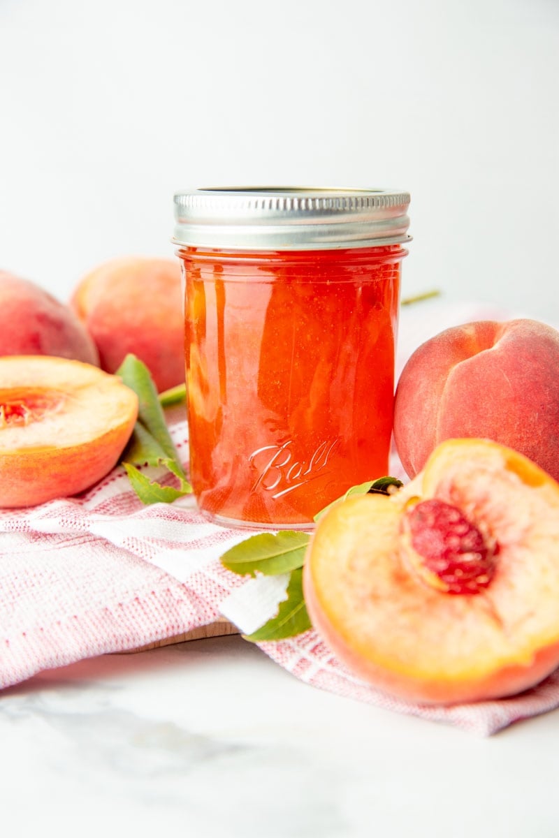 Bourbon peach jam sits in a half pint jar, surrounded by fresh peaches