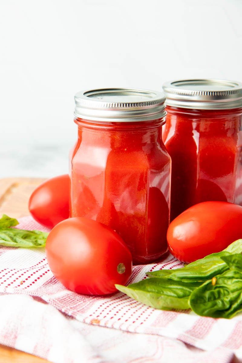 Close-up of two jars of canned basic tomato sauce with fresh roma tomatoes and basil around.