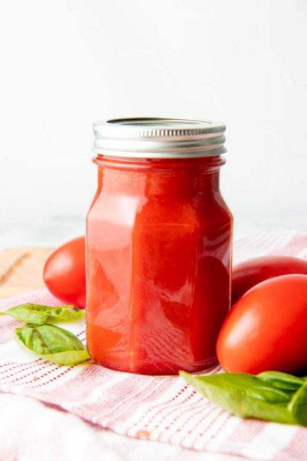 A single jar of canned tomato sauce stands surrounded by fresh tomatoes and basil.