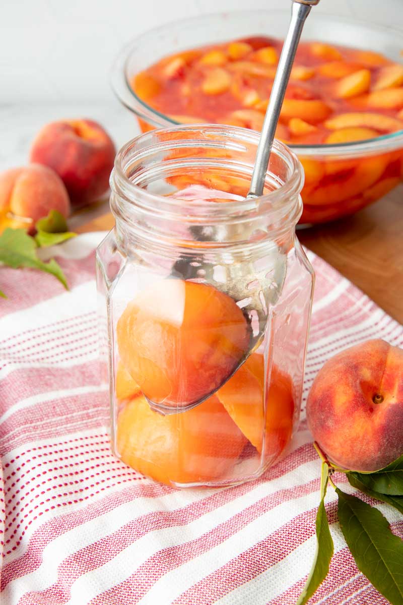 A slotted spoon lowers peach halves into a canning jar.