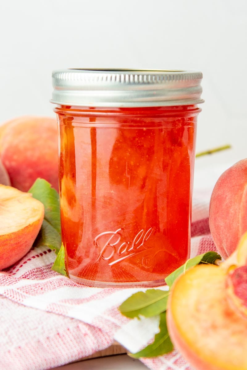 A jar of peach jam sits on a folded dishtowel, surrounded by fresh peaches