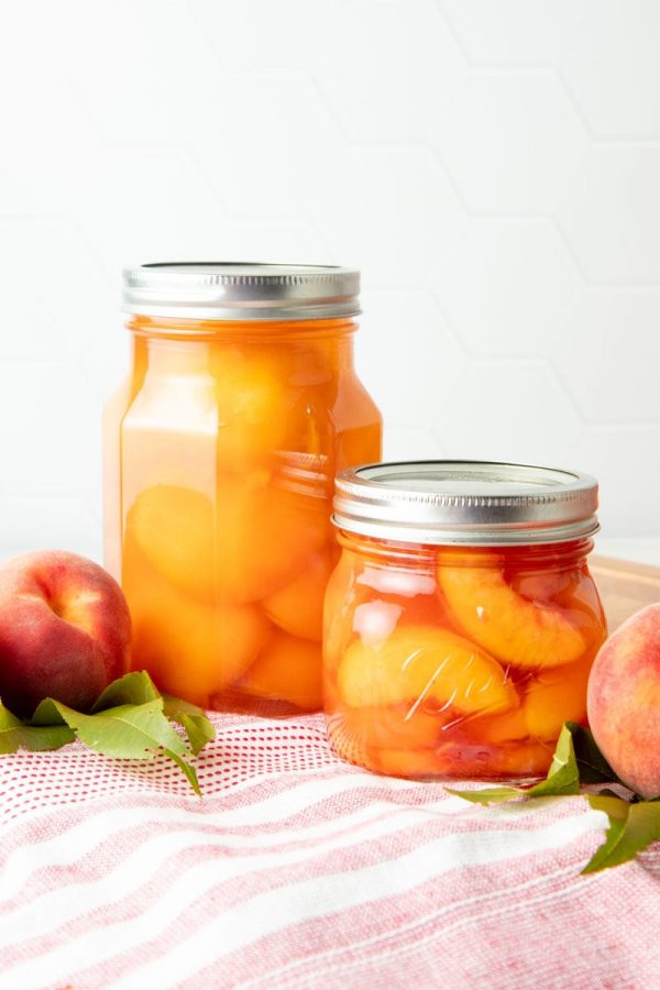 A pint jar of canned peach halves and a half-pint of canned peach slices stand side by side on a red and white kitchen linen.
