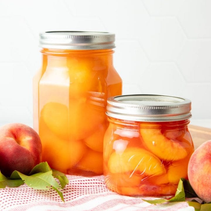 A pint jar of canned peach halves and a half-pint of canned peach slices stand side by side on a red and white kitchen linen.