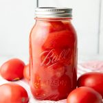 A jar of canned whole tomatoes sits atop a kitchen linen surround by fresh tomatoes.