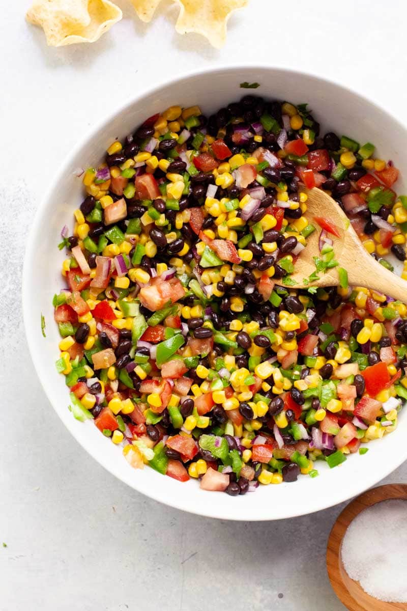 A wooden spoon scoops black bean and corn salsa from a large white serving bowl.
