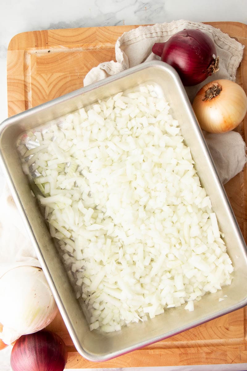 Overhead of a single layer of chopped onions on a cookie sheet with whole onions around it.