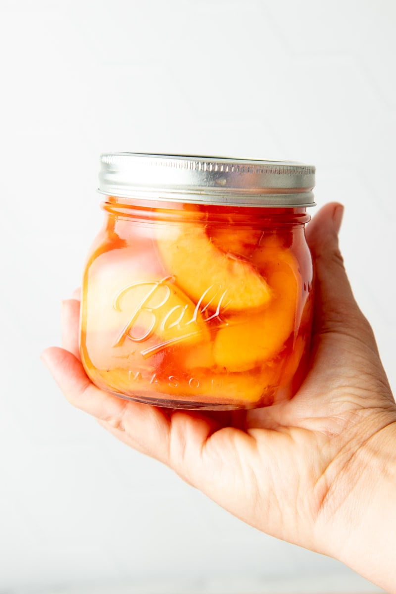 A hand holds up a small jar of canned peach slices.