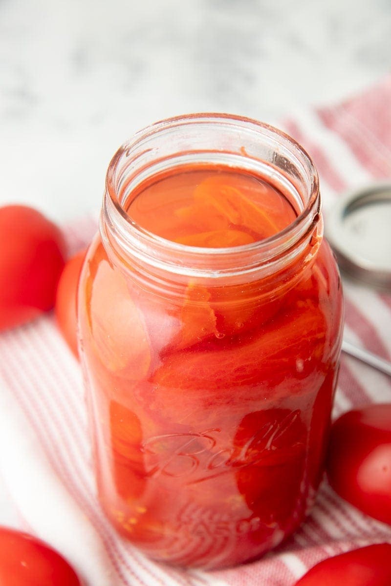 An open jar of whole tomatoes stands on a red and white dish towel.
