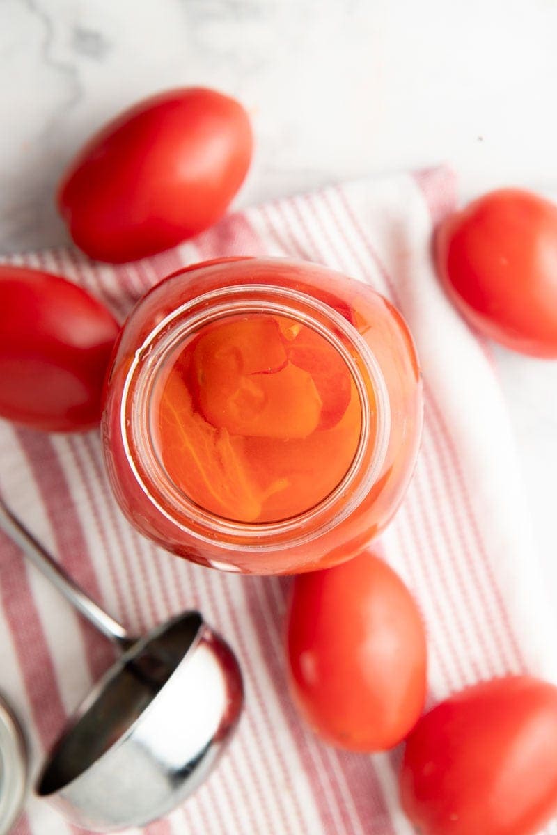 Overhead of tomatoes floating in juice in a canning jar, with a ladle and fresh tomatoes nearby.