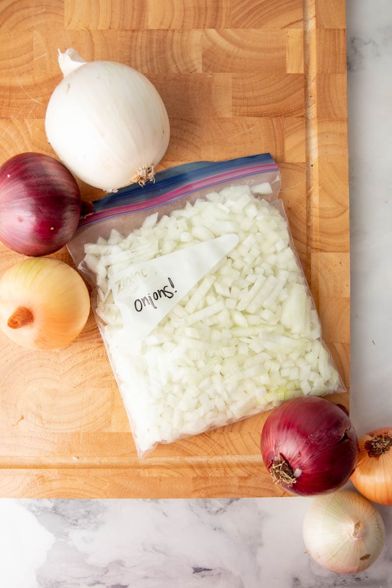 A Ziplock freezer bag of frozen onions rests on a wooden cutting board with whole onions around it.