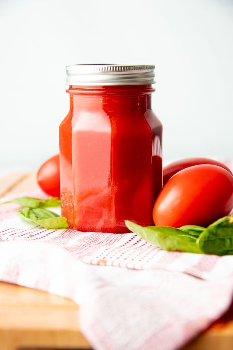 A closed jar of basic tomato sauce sits atop a kitchen linen with fresh ingredients around.