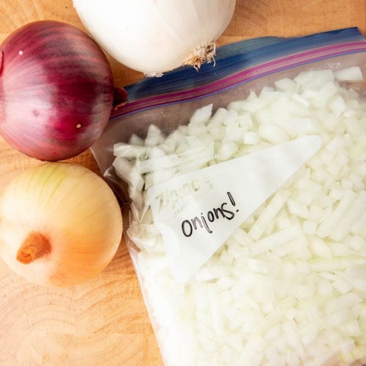 Close-up of a freezer bag filled with diced onions on a cutting board with three whole onions beside it.