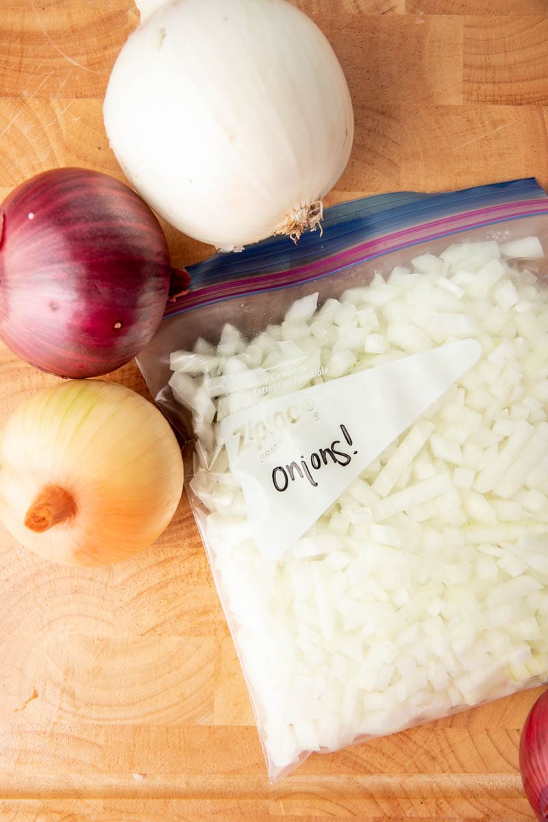 Close-up of a freezer bag filled with diced onions on a cutting board with three whole onions beside it.