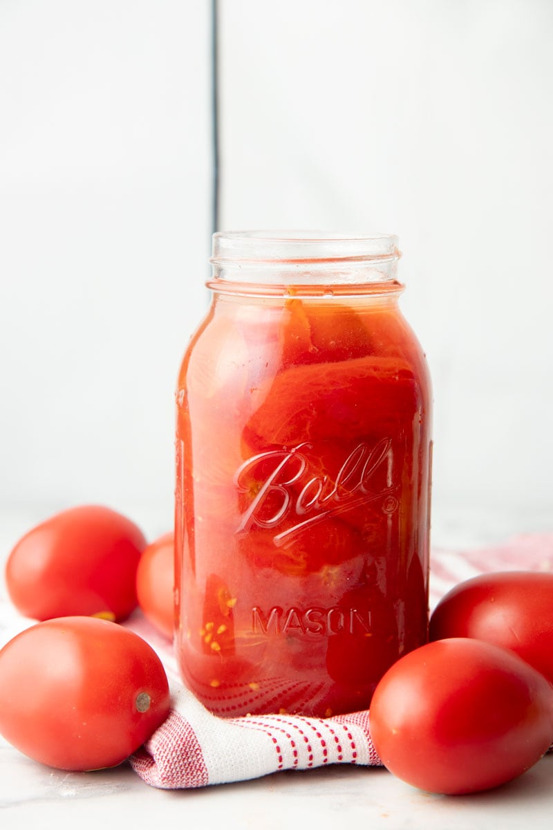 An open jar of canned whole tomatoes stands on a folded dish towel surrounded by fresh roma tomatoes.