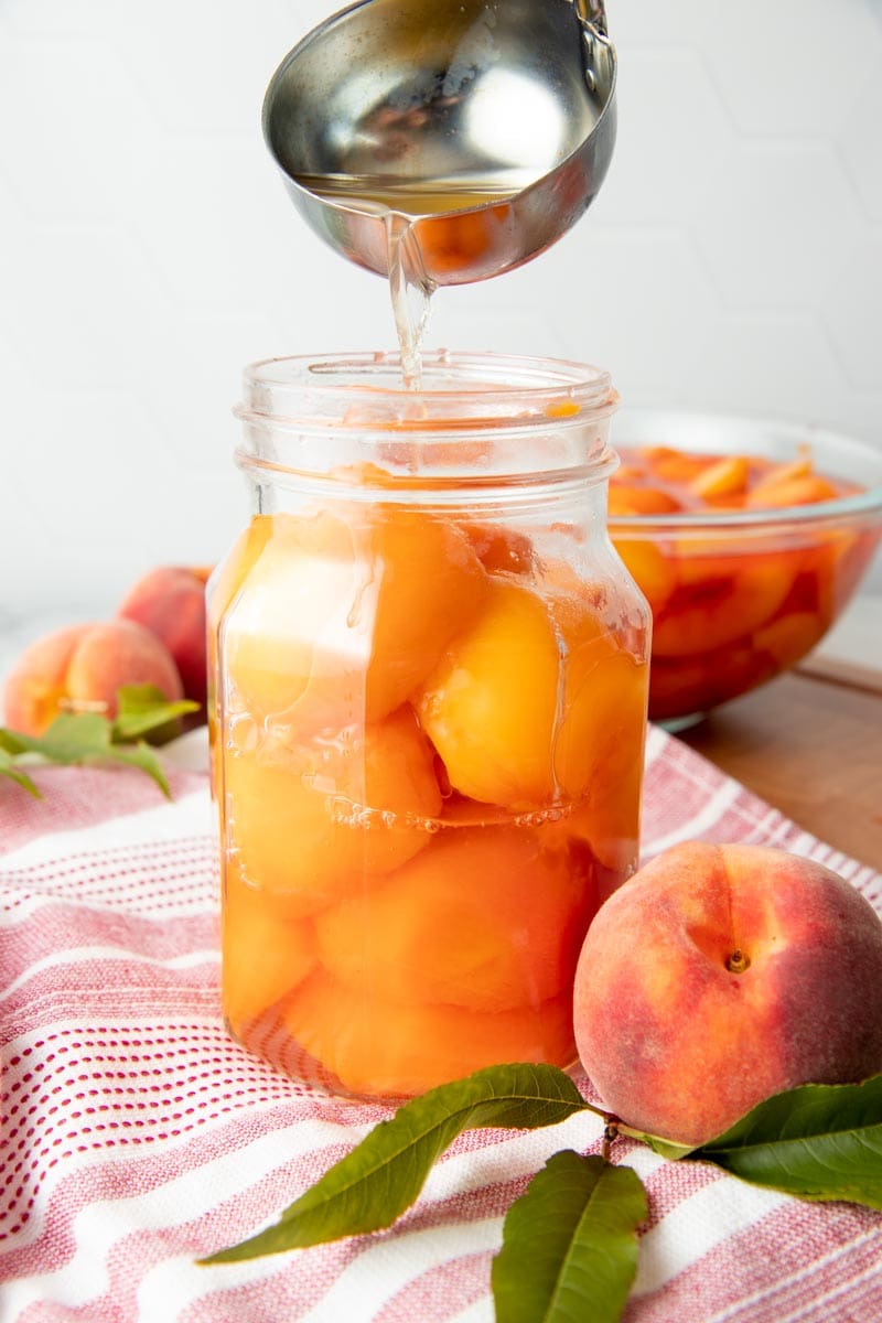 A ladle pours syrup into a jar of peach halves for canning.