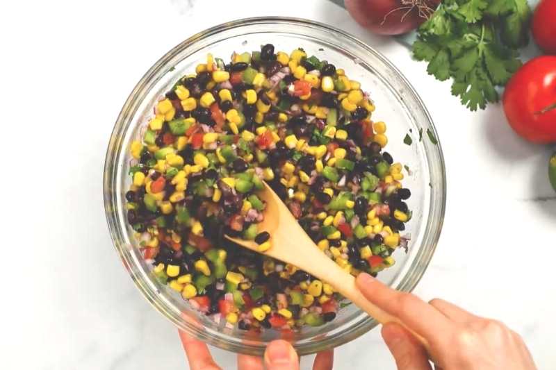 A wooden spoon mixes the salsa ingredients together in a glass bowl.