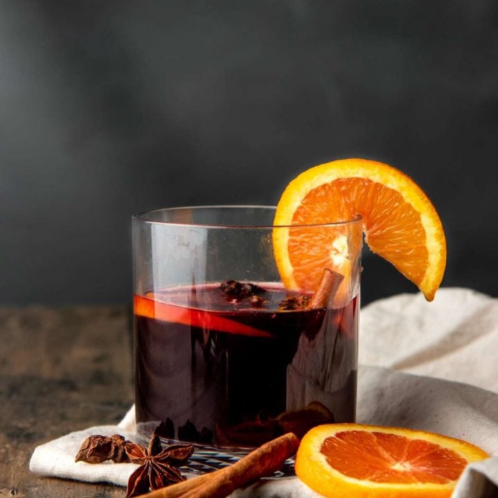 A glass of fall sangria sits on top of a white kitchen linen on a wooden table with whole spices and orange slices.