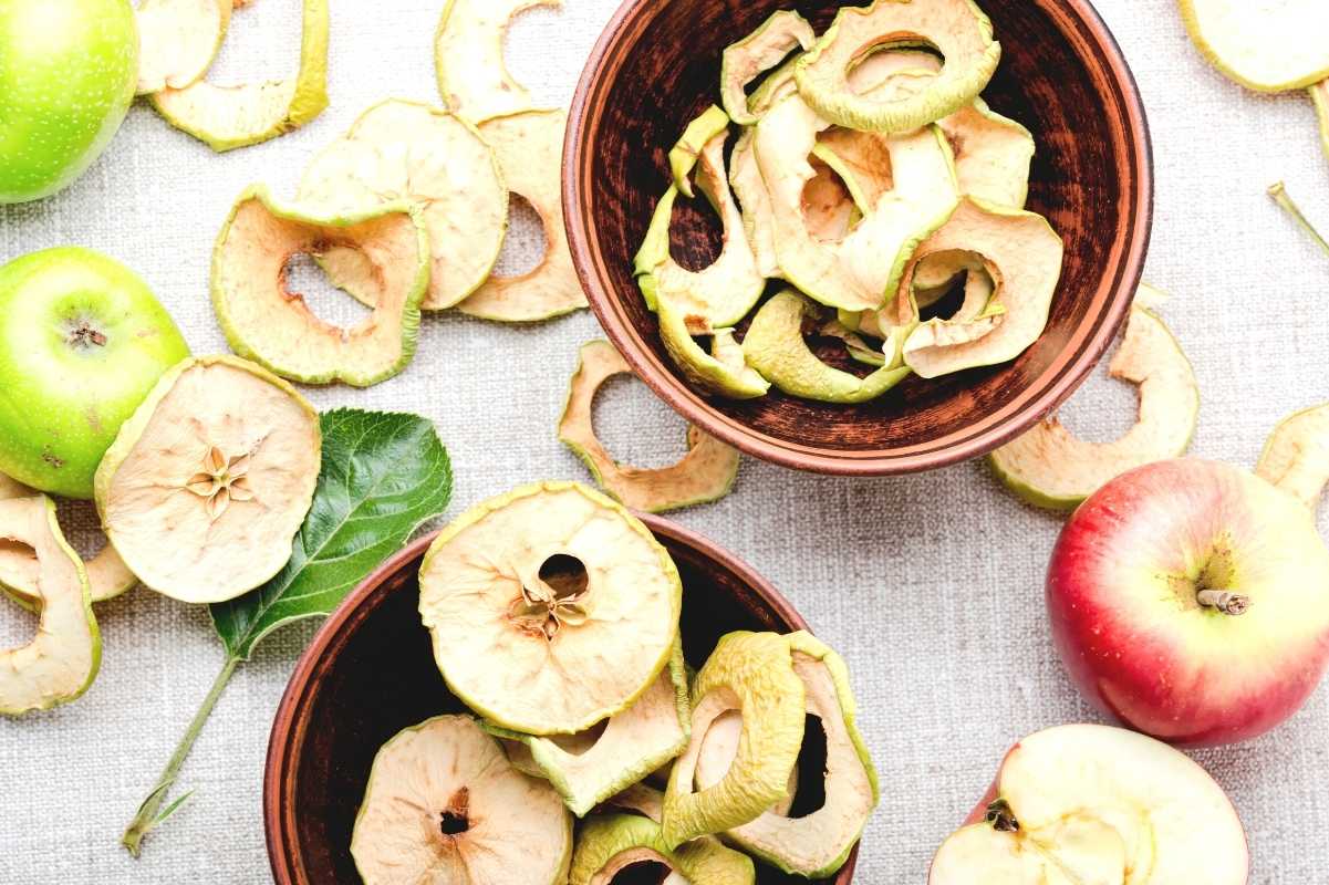 Close up on dried apple slices in two bowls with fresh apples and more slices surrounding them.