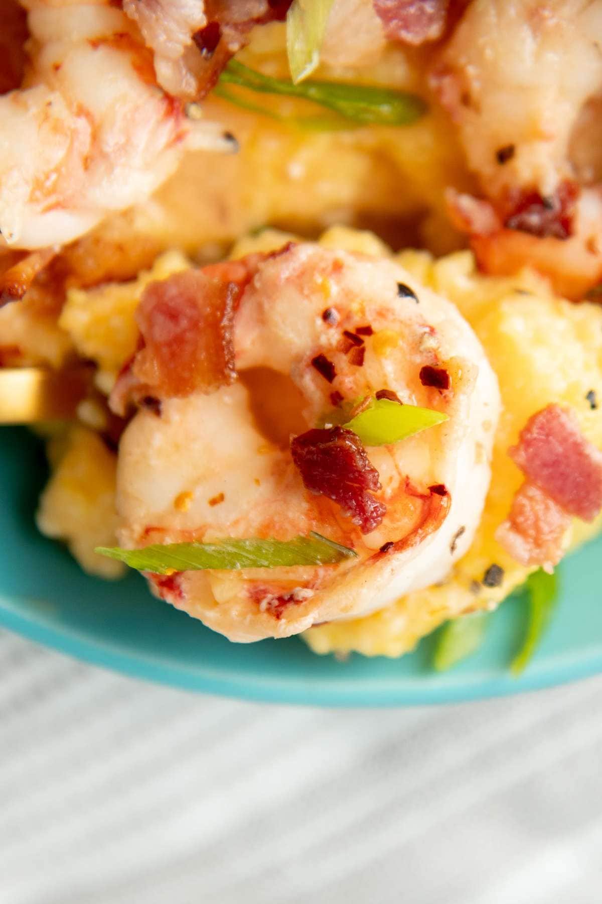 Close up of a plump, pink shrimp with crumbled bacon, red pepper flakes, and sliced green onion.