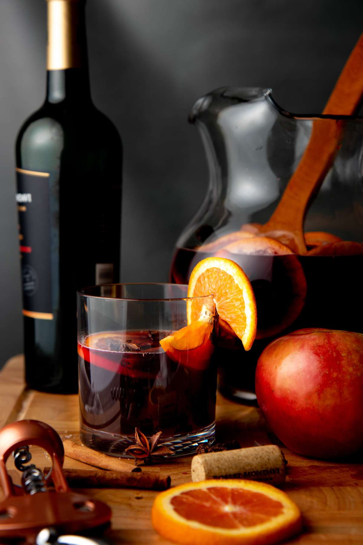 A rocks glass of apple cider sangria sits on a wooden cutting board in front of a bottle of red wine and pitcher of sangria.