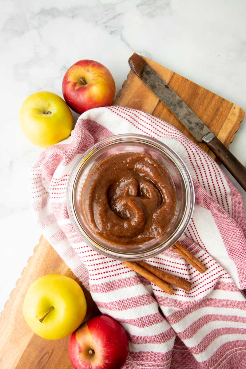 A jar of finished apple butter stands with cinnamon sticks and whole apples around it.