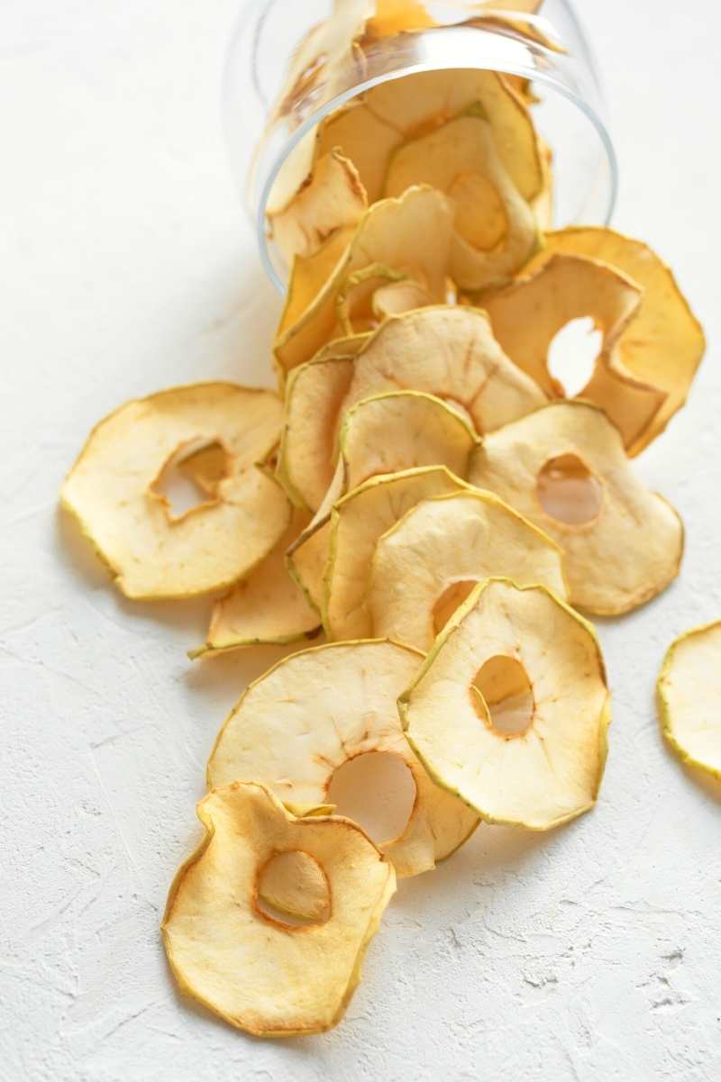 A glass jar spills out dried apple chips across a white counter.
