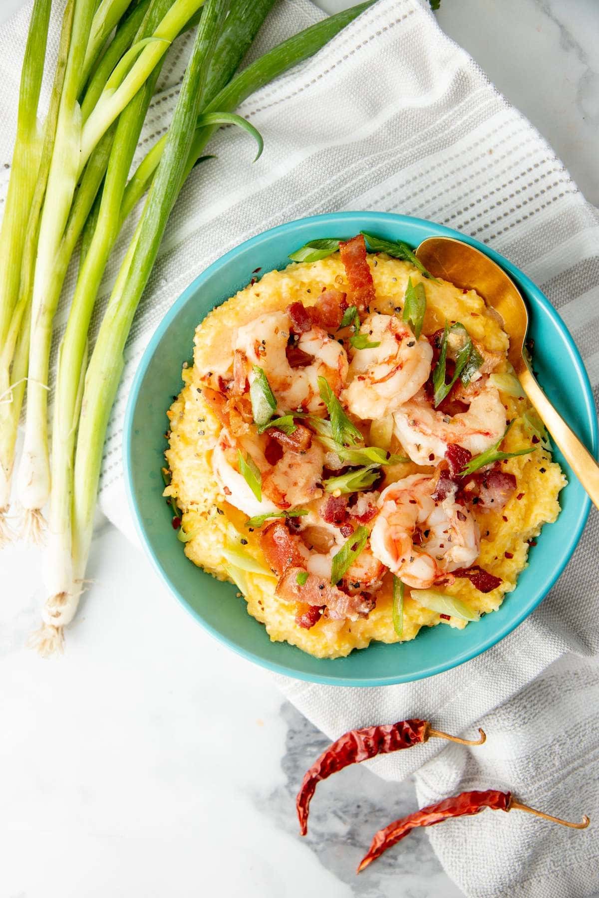 Top view of a bowl of grits topped with shrimp, bacon, and green onion with fresh scallions and dried chilies around it.