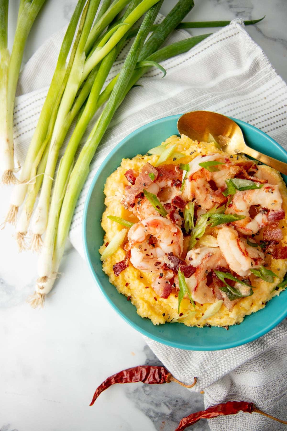 Overhead of a bowl of shrimp and grits on a kitchen towel with whole green onions and dried red peppers nearby.